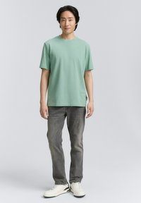 Camiseta de manga corta verde claro en tela suave, combinada con jeans gris y zapatillas blancas. Ajuste simple y relajado con escote redondo.