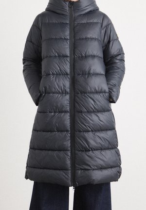 Persoon draagt een lange, zwarte, doorgestikte donzen jas met een ritssluiting aan de voorkant en een hoge kraag, handen in de zijzakken gestopt.