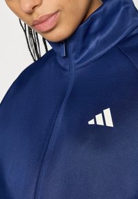 Tamsiai mėlynas striukės tipas su užtrauktuku, pagamintas iš lygaus audinio; turi aukštą apykaklę ir baltą logotipą su trimis juostelėmis kairėje pusėje.