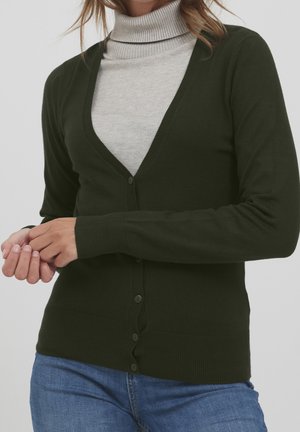 Femme portant un cardigan boutonné vert foncé sur un pull à col roulé gris clair et un jean bleu, les mains jointes devant elle.