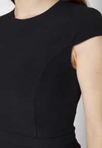 Robe noire avec un tissu structuré, des manches courtes et un corsage ajusté. Présente des lignes épurées et un détail de couture discret près de l'emmanchure.