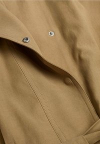 Gros plan sur un tissu marron clair avec deux boutons-pression argentés, montrant les coutures détaillées et la texture du tissu plié.