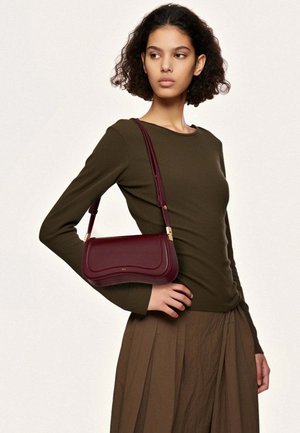 Femme portant un haut à manches longues olive et une jupe plissée marron, portant un sac bandoulière en cuir bordeaux avec des accents dorés.