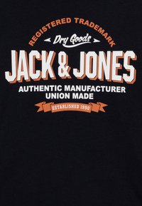 Crna pamučna majica s bijelim i narančastim grafičkim tekstom, koji uključuje "Jack & Jones", "Autentični proizvođač" i "Osnovano 1990."