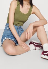 Olivgrön ribbad linne, högmidjade slitna denimshorts, vinröda sneakers med vita sulor och vita strumpor, i sittande position.