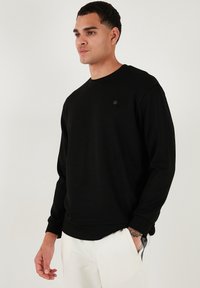 Sudadera negra de corte relajado, fabricada en tejido suave, con un cuello redondo de canalé y un discreto detalle del logo en el pecho.
