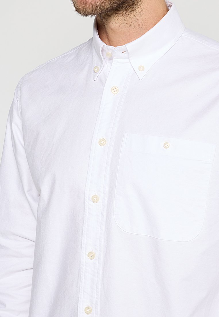 Man met een witte overhemd met knoopsluiting en een geknoopte kraag, en een enkele geknoopte borstzak, close-up view.