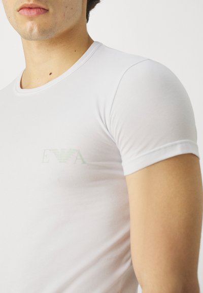 Camiseta blanca de algodón con mangas cortas, que presenta un sutil logotipo verde en el pecho. Textura suave y forma ajustada.