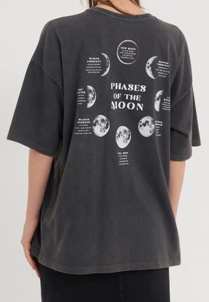 Person wearing a dark oversized t-shirt showing moon phases and corresponding descriptive text on the back.
