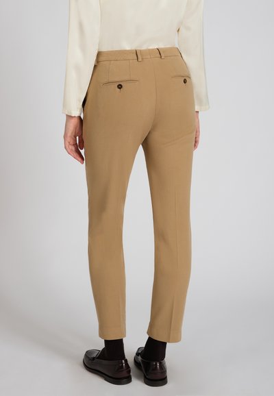 Beżowe spodnie o dopasowanym kroju, charakteryzujące się gładką teksturą, dwoma tylnymi kieszeniami i zamkiem błyskawicznym. Połączone z ciemnymi butami i skarpetkami.