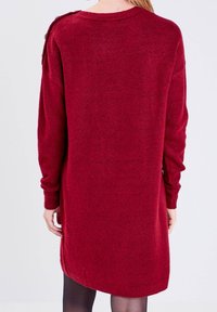 Vestido de punto burdeos con mangas largas, escote redondo y detalles con botones en los laterales. La parte trasera tiene un dobladillo ligeramente más largo. Tejido suave.