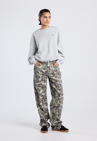 Persona con el cabello peinado hacia atrás, vistiendo un sudadera oversized de color gris claro, pantalones de camuflaje en verde y gris, y zapatillas negras, de pie con las manos en los bolsillos.