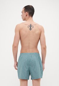 Shorts de bain rayés en teal avec des lignes blanches, taille élastique, longueur mi-cuisse. La vue arrière montre un tatouage de fleur de lis dans le bas du dos.