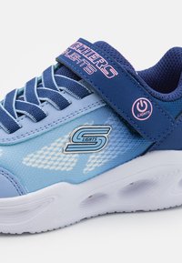 Plava sportska cipela s tekstilnim gornjim dijelom, mrežastim detaljima, čičak trakom i bijelim potplatom. Ima logo i svjetla sa strane.