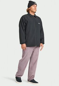 Homem a usar um gorro preto, uma jaqueta preta com fecho eclair, calças cor mauve e ténis pretos, em pé com uma postura relaxada em fundo branco.