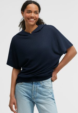 Frau mit lockigem Haar lächelt, trägt ein marineblaues, strukturiertes kurzärmeliges Oberteil und hellblaue Jeans vor schlichtem Hintergrund.