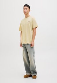 Tricou bej din bumbac, cu mâneci scurte și decolteu rotund, având un logo mic pe partea stângă a pieptului. Blugi ușor largi, de culoare albastru deschis.