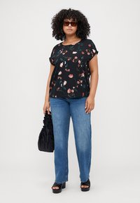 Black floral top with short sleeves over light blue straight-leg jeans. Model carries a black ruched handbag and wears platform sandals.