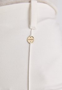 Weiße hochtaillierte Hosen mit schmalem Schnitt, strukturierter Stoff und einem goldenen, runden Logo-Detail auf der Vorderseite in der Nähe des Bundes.