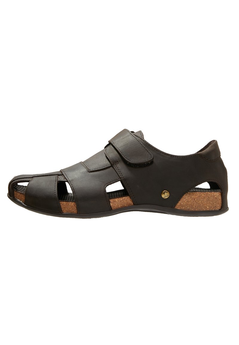 Black leather sandal with open cutouts, a velcro strap, cork insole, and a rubber outsole. Features a rounded toe design and minimal hardware.
