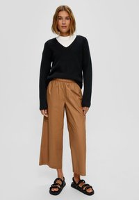 Zwarte gebreide trui met een V-hals, gecombineerd met losse, bruine cropped broeken en zwarte sandalen met bandjes, die eenvoud en comfort benadrukken.