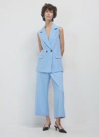Gilet su misura azzurro chiaro e pantaloni a gamba larga, con scollatura a V profonda, chiusura frontale a doppio bottone e mules neri con tacco. Texture liscia.