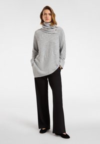 Grijze kabelgebreide trui met een hoge kraag en knoopdetail, gecombineerd met zwarte wijde broek en zwarte platte schoenen.