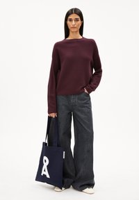 Kastanienbrauner strukturierter Pullover, weit geschnittene graue Jeans und eine marineblaue Tasche mit einem weißen grafischen Design. Das Model steht vor einem schlichten Hintergrund.