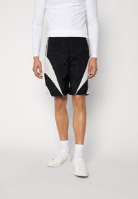 Person som bär svarta och vita idrottsshorts, vit långärmad tröja, vita strumpor och vita sneakers, stående mot en ljus bakgrund.
