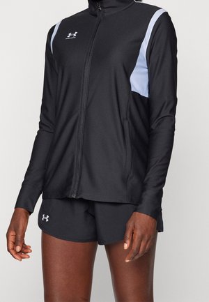 Czarna kurtka na zamek firmy Under Armour z białymi akcentami na ramionach, noszona z czarnymi szortami, ukazująca ramiona i górną część nóg stojącej osoby.