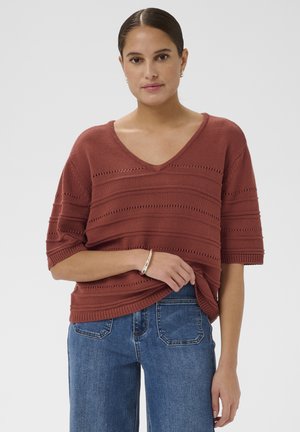 Vrouw die een roestkleurige V-hals gebreide trui met korte mouwen en een blauwe spijkerbroek draagt, staande tegen een effen lichte achtergrond.