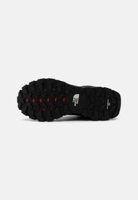 Suela de goma negra con un patrón de banda de rodadura agresivo y acentos rojos; presenta el logo de The North Face embossado en el lateral.