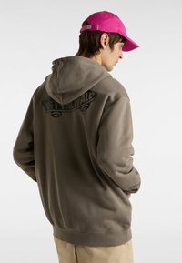 Brauner Kapuzenpullover mit einem großen schwarzen "OFF THE WALL"-Grafik auf dem Rücken, aus Baumwollmaterial, lockerer Schnitt, getragen mit einer pinken Mütze.