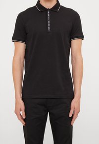 Polo noir en coton, avec un col et des poignets contrastants en blanc, affichant un détail de logo vertical sur la patte de boutonnage.