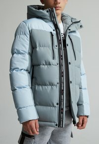 Veste matelassée bleu clair avec fermeture éclair à l'avant, accents de fermeture éclair noire, deux poches sur la poitrine et une capuche. Présente des détails de logo le long de la fermeture éclair.