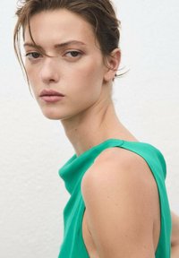 Jonge vrouw met kort haar in een mouwloze groene top, die iets opzij kijkt tegen een eenvoudige lichte achtergrond.