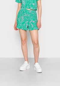 Calções verdes com padrão, apresentando uma bainha franzida e um design floral em rosa e branco, combinados com ténis brancos de sola espessa.