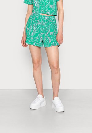 Calções verdes com padrão, apresentando uma bainha franzida e um design floral em rosa e branco, combinados com ténis brancos de sola espessa.