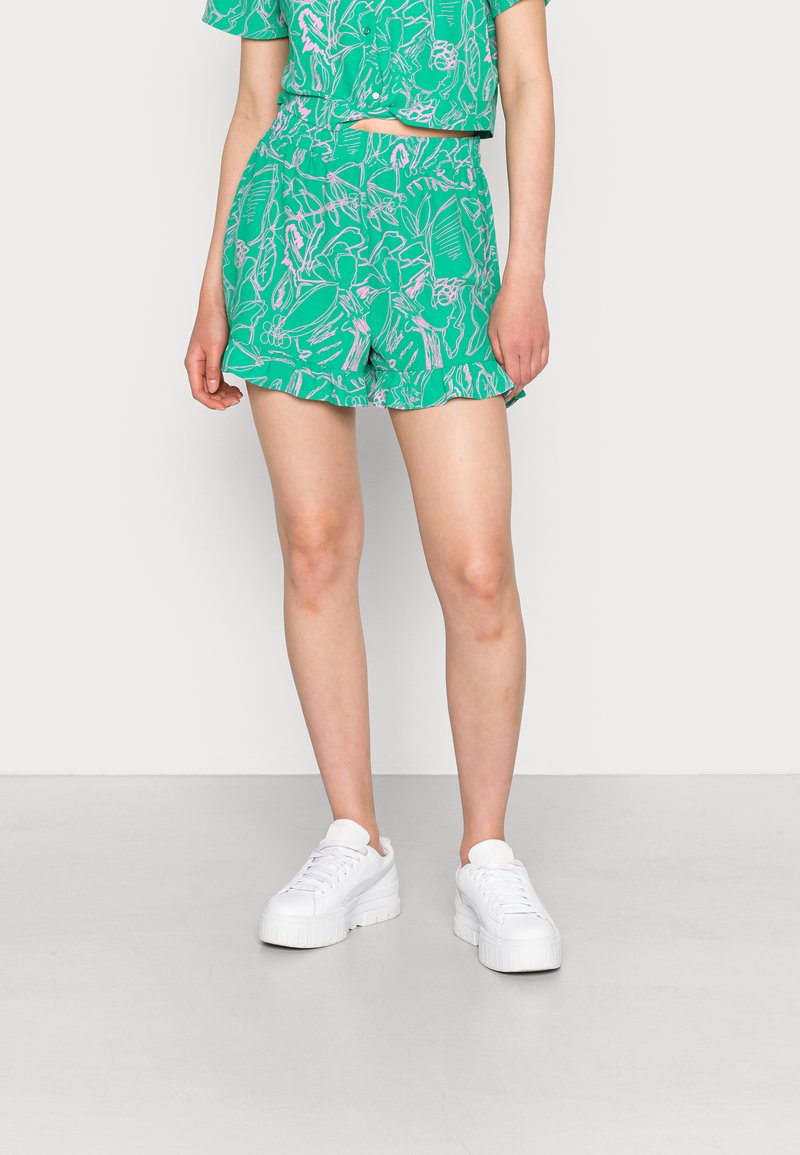 Calções verdes com padrão, apresentando uma bainha franzida e um design floral em rosa e branco, combinados com ténis brancos de sola espessa.