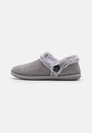 Chausson gris à enfiler avec une doublure douce en fausse fourrure et un bouton décoratif sur le côté, doté d'un extérieur imitation daim et d'une semelle cousue.
