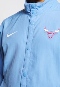 Ljusblå Nike-jacka med hög krage, tryckknappsstängning och en röd och vit tjur-logotyp på vänster bröst. Slät, lättviktsmaterial.