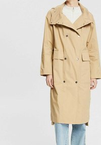 Cappotto trench lungo beige con bottoni doppi in argento, tasche a palla, indossato sopra un maglione bianco e jeans azzurri chiari.