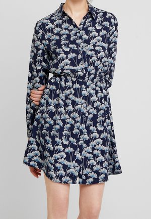 Woman wearing a navy blue, long-sleeve shirt dress with white and light blue floral print and a cinched waist.