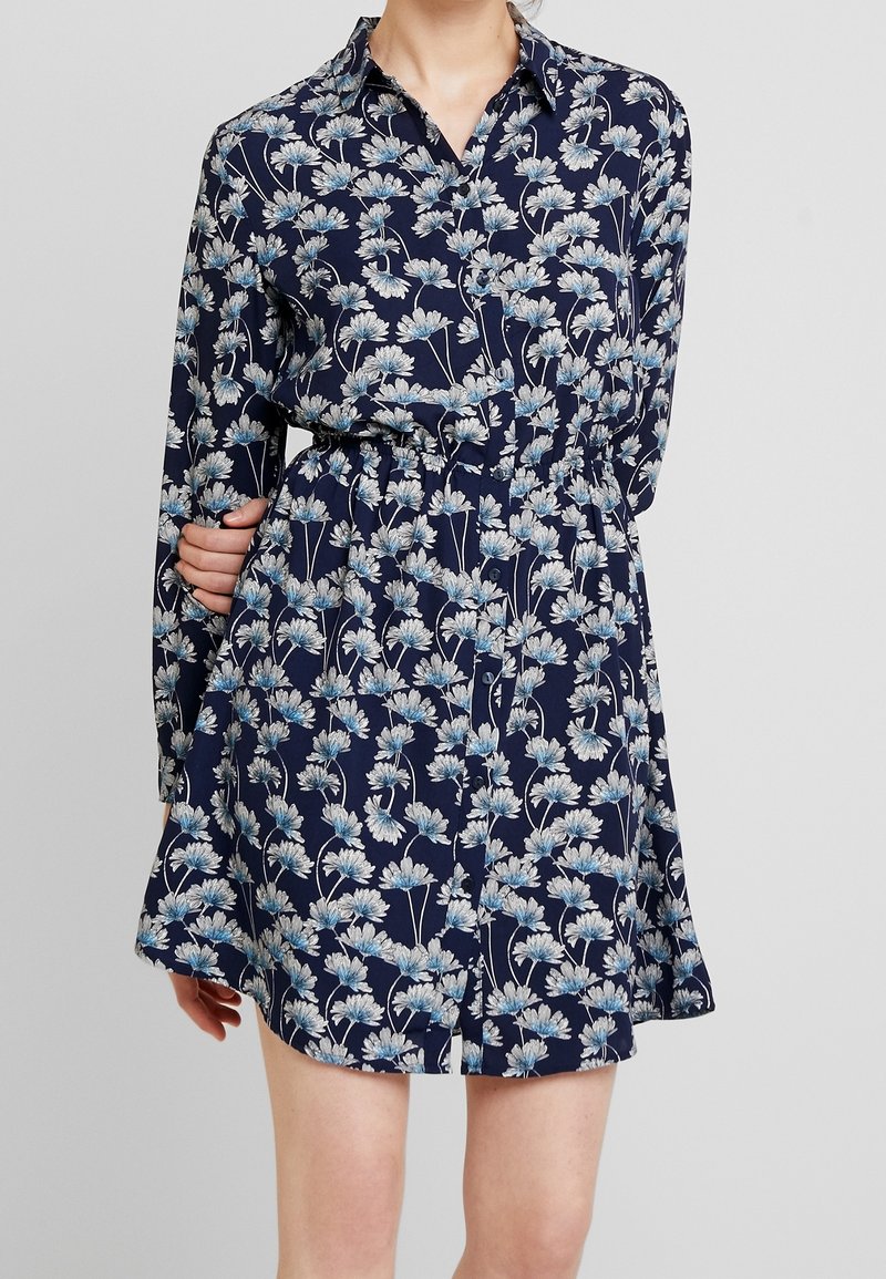 Femme portant une robe-chemise bleu marine à manches longues avec un imprimé floral blanc et bleu clair et une taille cintrée.