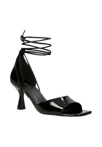 Sandalo con tacco alto in vernice nera, punta aperta, suola anteriore quadrata e cinturini avvolgenti alla caviglia su sfondo bianco.