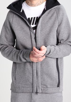 Personne portant un sweat à capuche gris zippé sur un t-shirt blanc avec le logo Nike, les mains jointes devant et des tatouages visibles au niveau du cou.