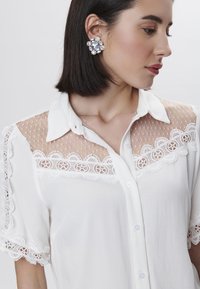 Blusa bianca con rete trasparente e dettagli in pizzo ricamato sulle spalle e sull'orlo. Il modello indossa un grande orecchino gioiello.