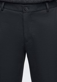 Κοντινό πλάνο μαύρου υφασμάτινου παντελονιού με κούμπωμα με κουμπιά και θηλιές για ζώνη, φορεμένο με σκοτεινή μπλούζα που έχει μπει μέσα.