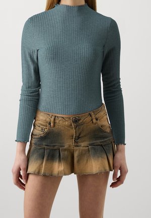 Femme portant un haut côtelé bleu sarcelle ajusté à manches longues et une mini-jupe plissée marron et noire délavée, debout devant un fond uni.