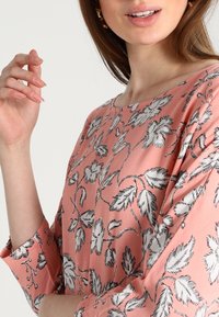 Rosa blus med svarta blommönster, lös passform, 3/4-långa ärmar, slät textur, rund halsringning och subtil lyster.